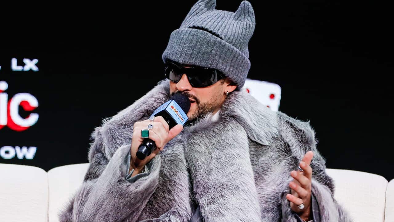 Bad Bunny holds a microphone to his mouth, sitting on a couch wearing a grey fur jacket and beanie with cat ears.
