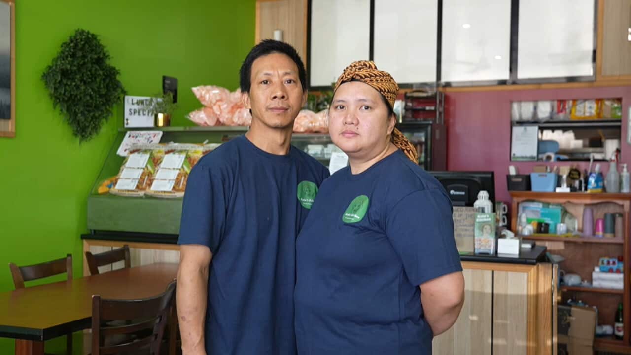 A couple wearing matching navy shirts stand side by side inside a small restaurant, with a food display and counter behind them.