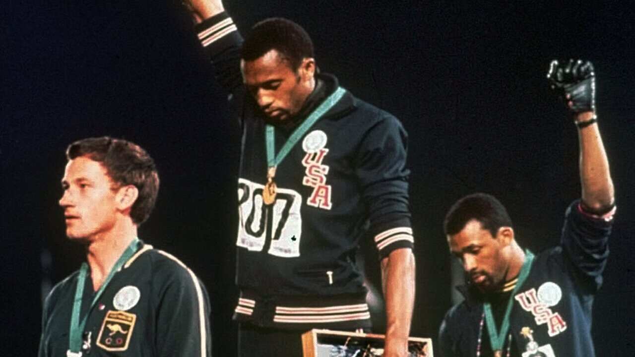 At the 1968 games, Australian silver medalist Peter Norman stands beside US Athletes Tommie Smith and John Carlos as they raise their gloved hands in protest.