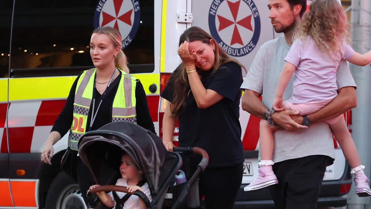 A woman rubs tears from her eyes as she pushes a pram, flanked by a police officer on one side and a man holding a small child in the other. Ambulance in the background.