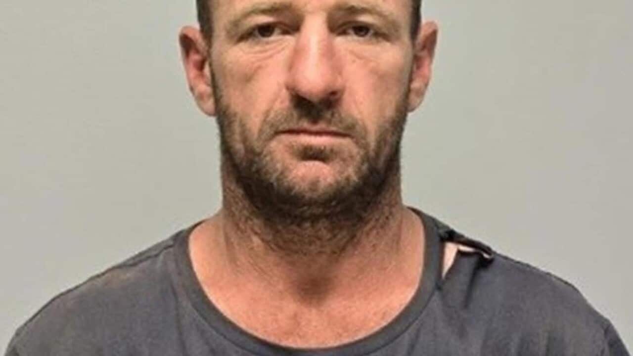A head-and-shoulders portrait of a middle-aged man with short dark hair, light stubble, and a small visible injury on his forehead, wearing a worn grey t-shirt against a plain neutral background.