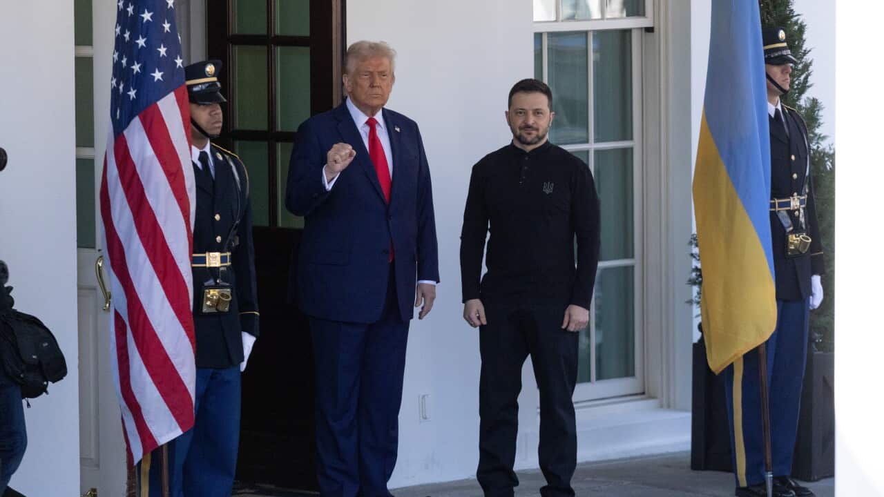 A man wearing a suit and tie stands beside another man wearing military fatigue with two guards standing nearby.