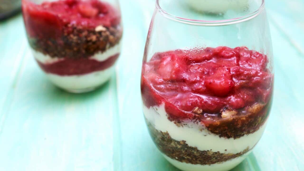 Two glasses sit on an greenish table top. Each glass holds layers of yoghurt and granola, topped with vibrant pink stewed fruit.