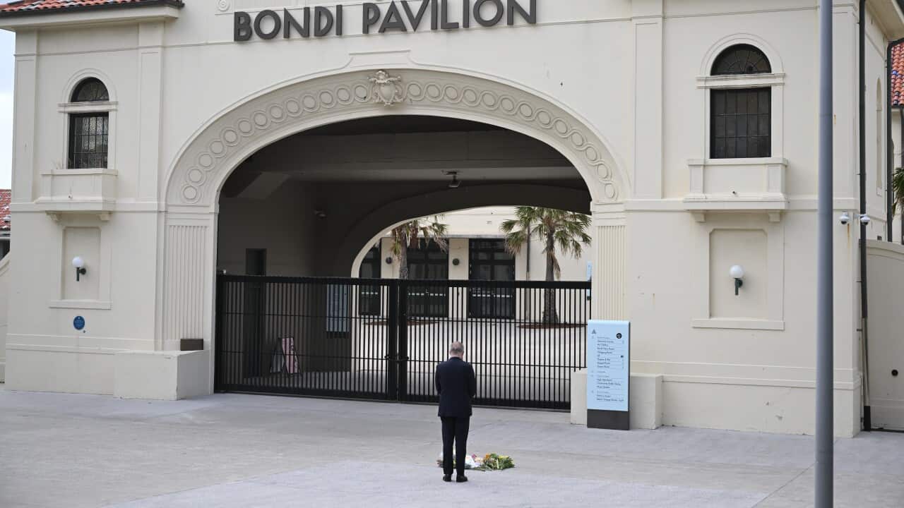A man dressed in a black suit stands in front of a gate with flowers at his feet.