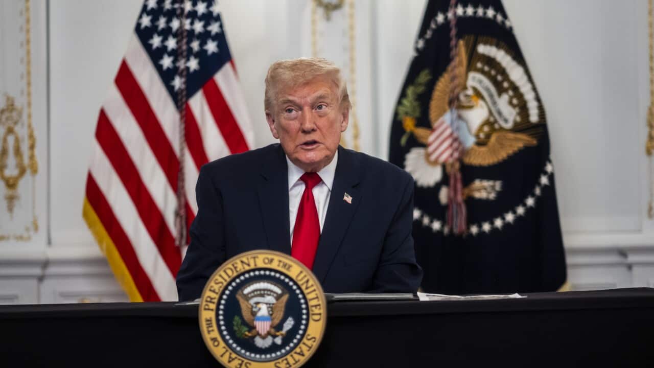 US president Donald Trump at a table with the US and a military flag behind him