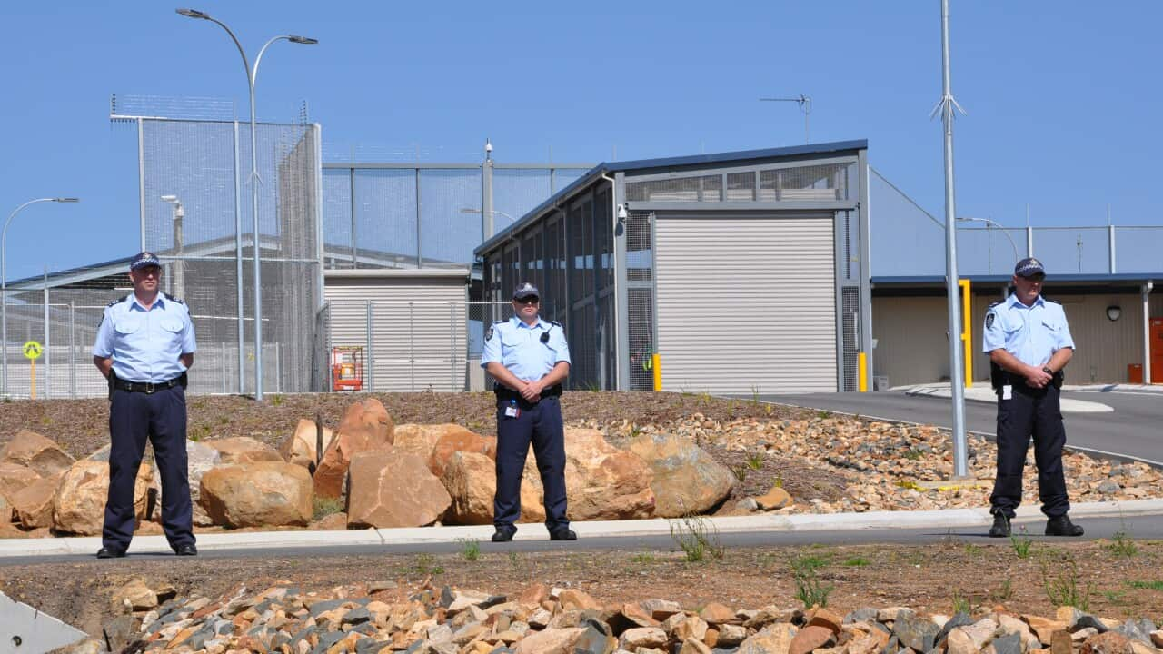 Three police officers stand in front of what appears to be a high-security compound in an arid environment.