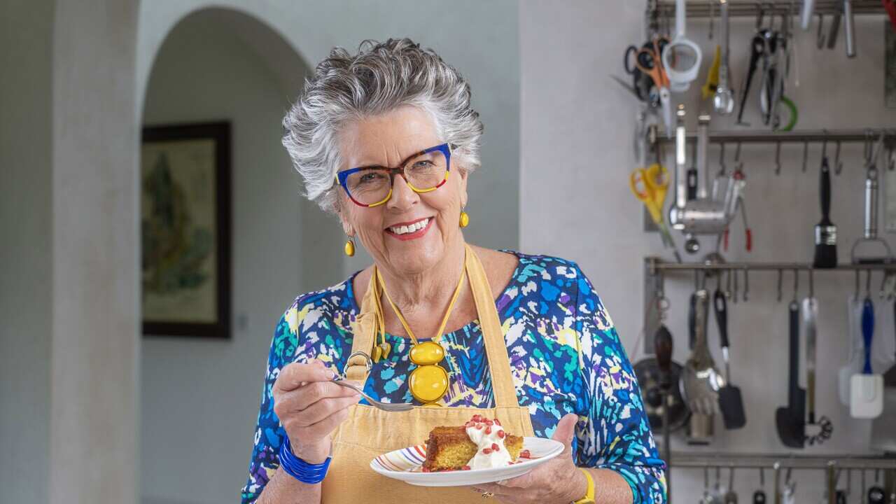 Prue Leith, in a brightly patterned blue dress and yellow apron, stands in her kitchen, holding a plate with cake and cream.