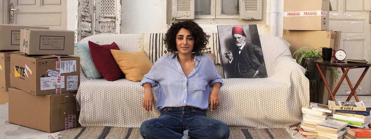 A woman in blue jeans and a blue shirt sits on a rug on the floor, in front of a couch. Packing boxes are stacked to either side of the couch. Several piles of books sit on one side.