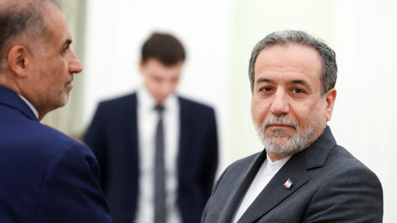 A man wearing a suit looks at the camera with another man standing in front of him.