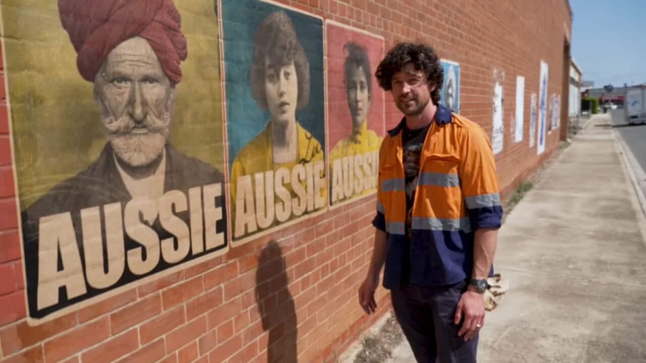 Artist Peter Drew stands by three posters wearing an orange hi-vis shirt. The posters show three people of different ethnicities with the text AUSSIE emblazoned below.