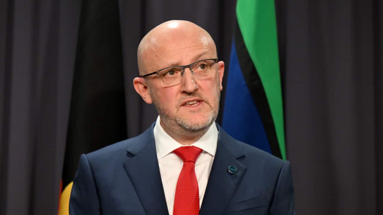 A man in a dark suit with a red tie stands in front of a black background