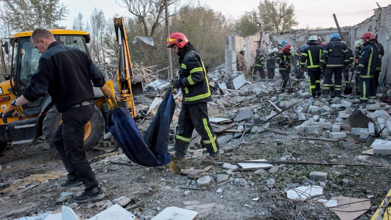 Man in personal protective equipment and a helmet holds tarp with another man, as they walk through building rubble. A group of other people in PPE gather together for cleanup efforts.