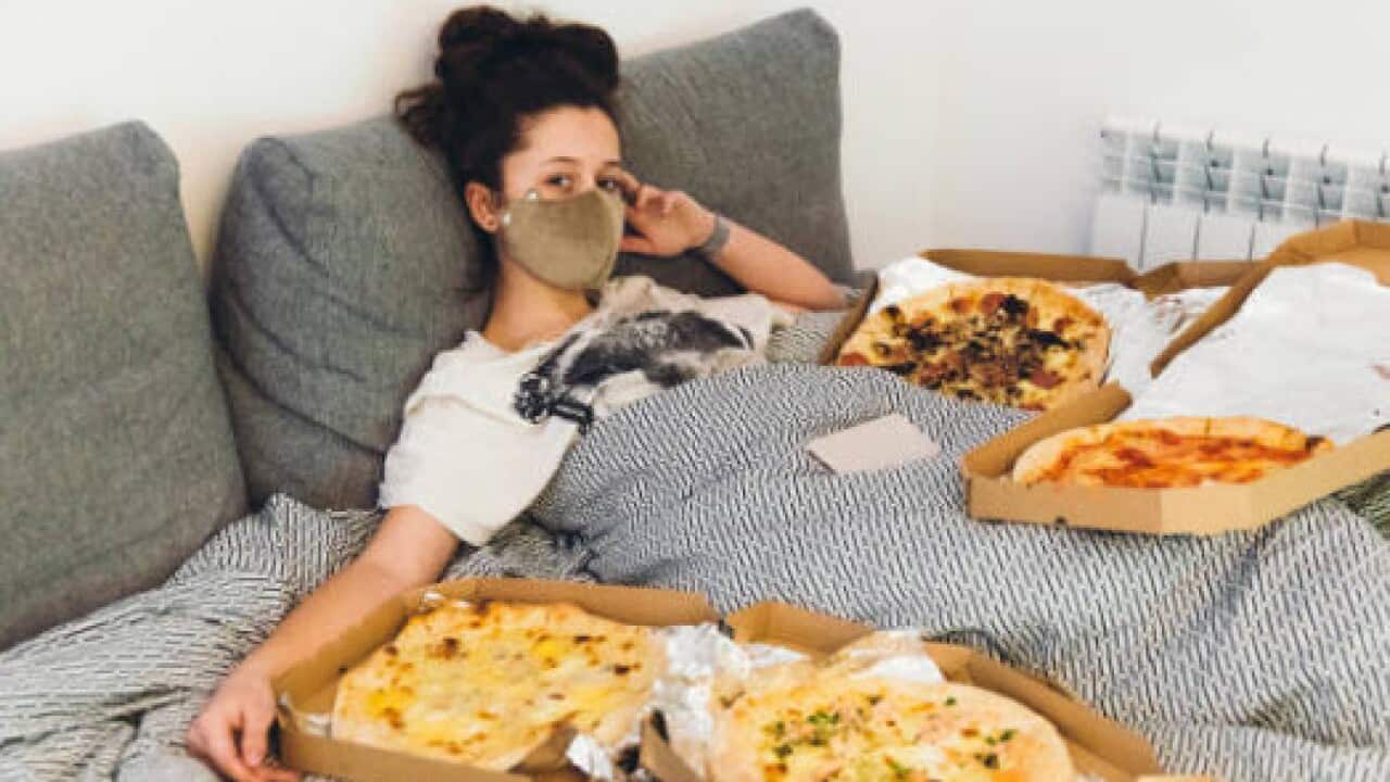 Young woman wearing a protection mask from coronavirus lying in bed surrounded by pizzas at her apartment.