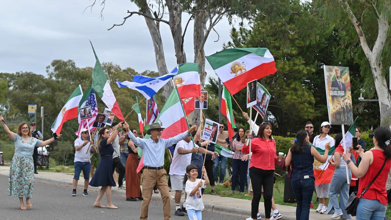 A group of demonstrators marches down a street carrying American flags and Iranian Lion and Sun flags alongside posters featuring Rezā Pahlavi.