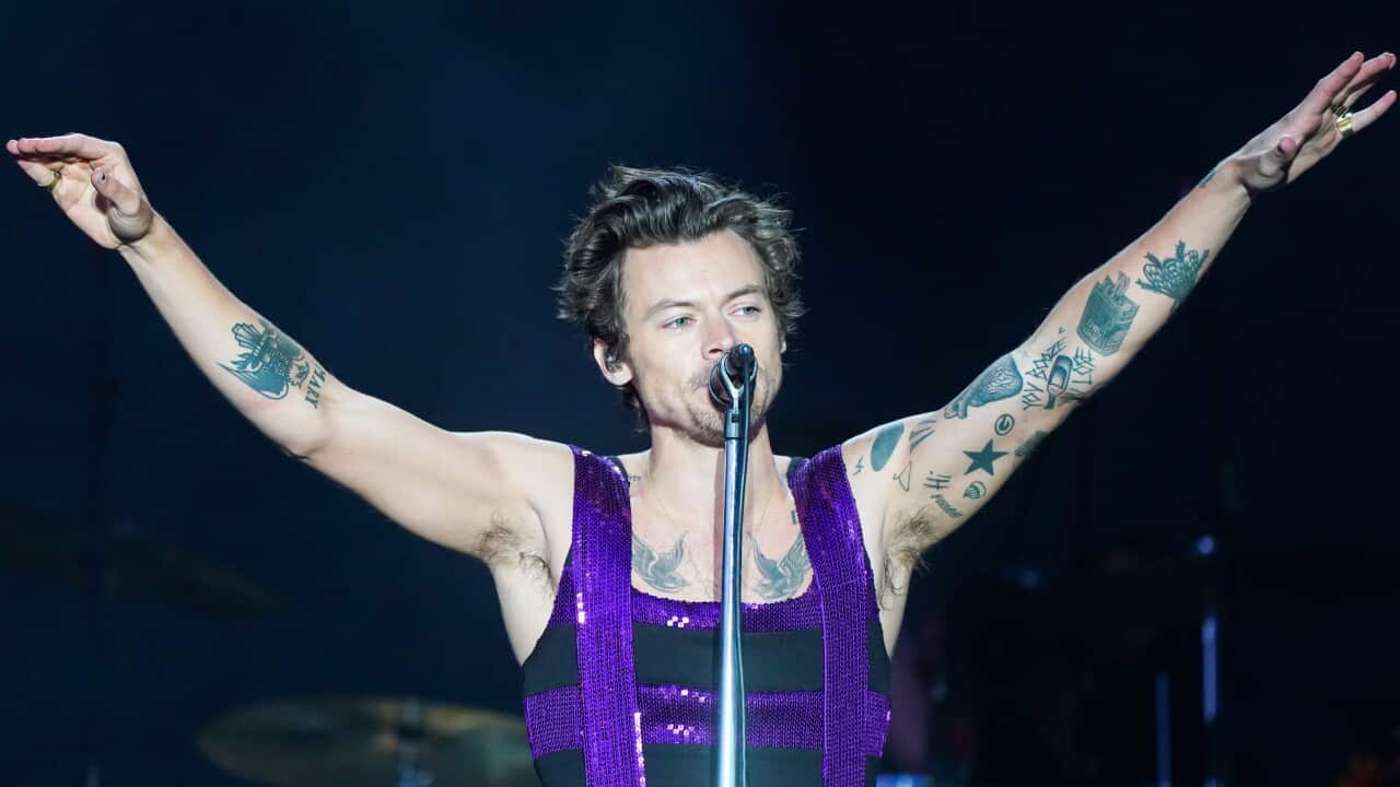 Harry Styles singing into a microphone. He is wearing a purple and black jumpsuit.