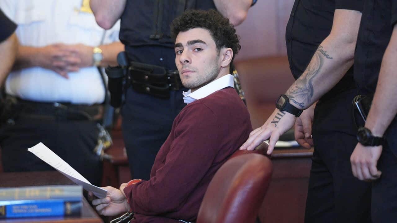 A young man with short curly black hair sits at a wooden desk holding several sheets of paper in his right hand.