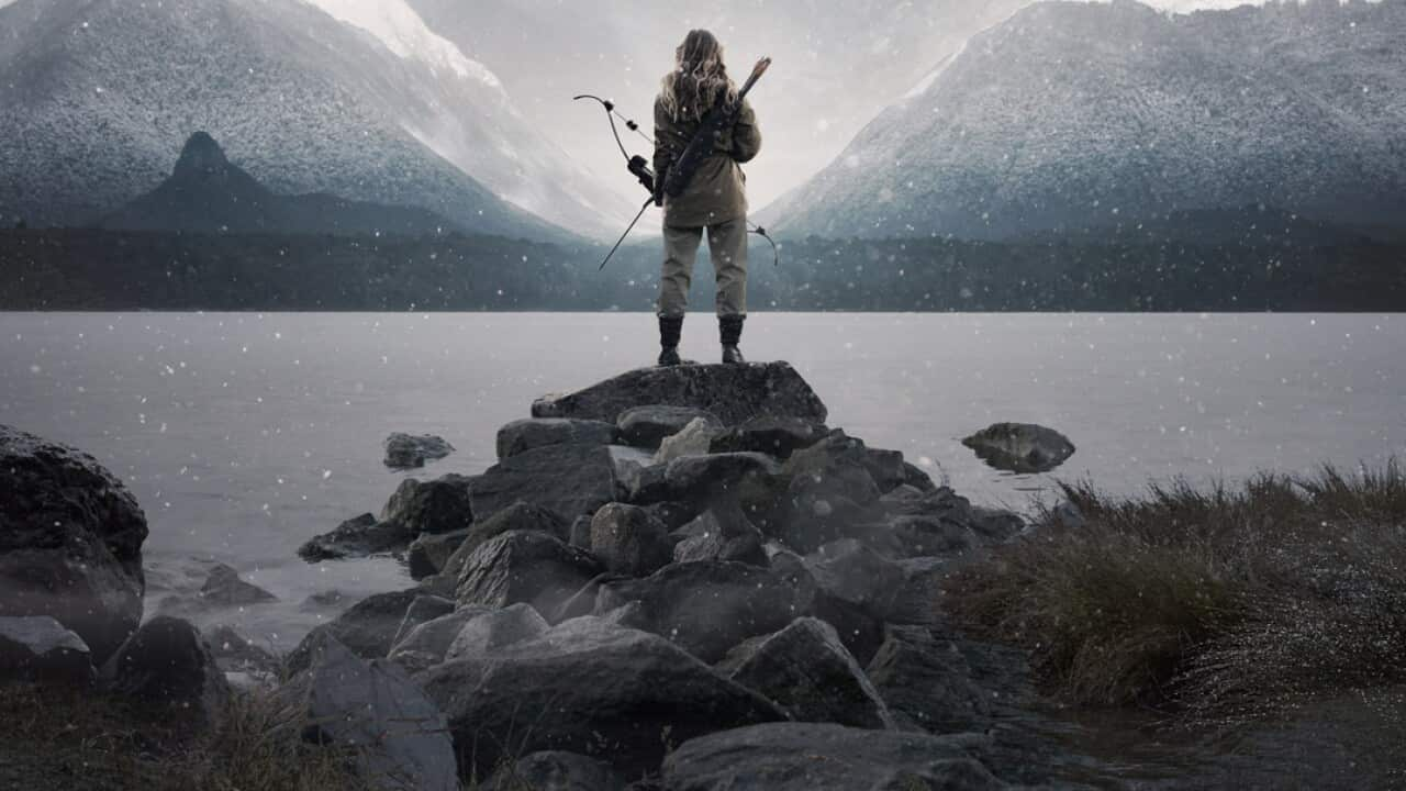 A person holding a bow and arrow is seen from behind, standing by a sweeping body of water. The image is in tones of grey, with snow-capped mountains in the distance. 