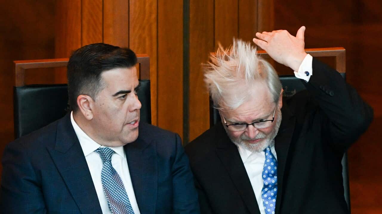 Kevin Rudd and another man sit wearing suits. Rudd is adjusting his hair.