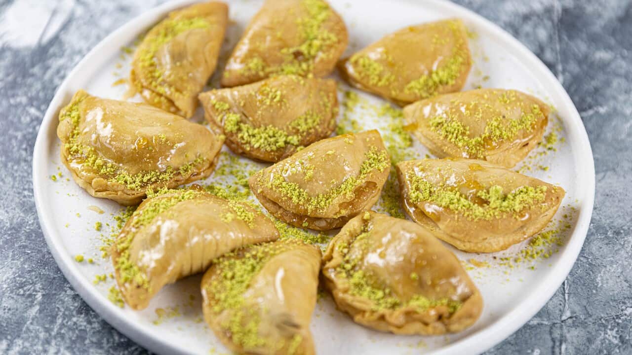 A round white plate holds 10 half-moon shaped small golden pastries. Each is sprinkled with crushed pistachios.