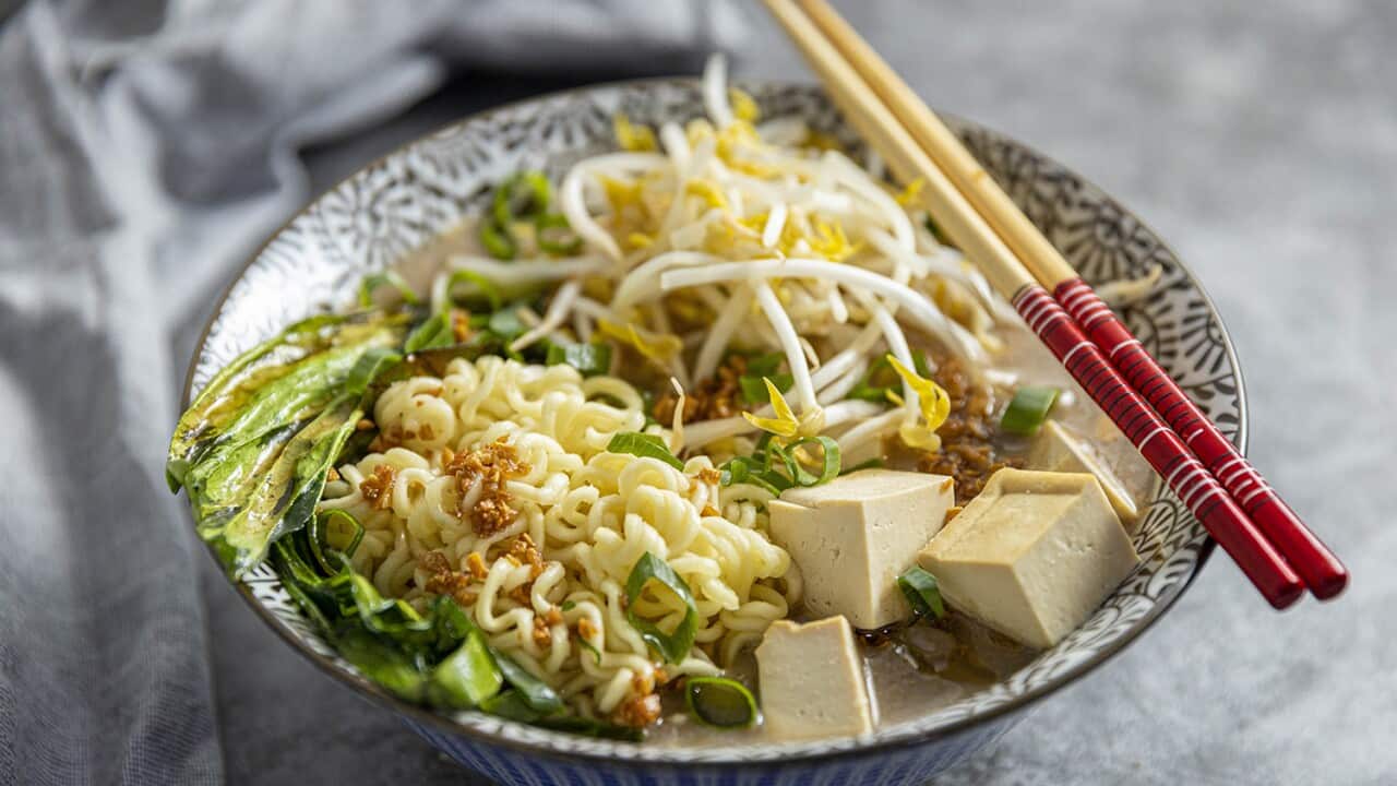 Chopsticks rest across one side of a bowl. The bowl has liquid at the bottom, topped with noodes, cubes of tofu and other toppings.
