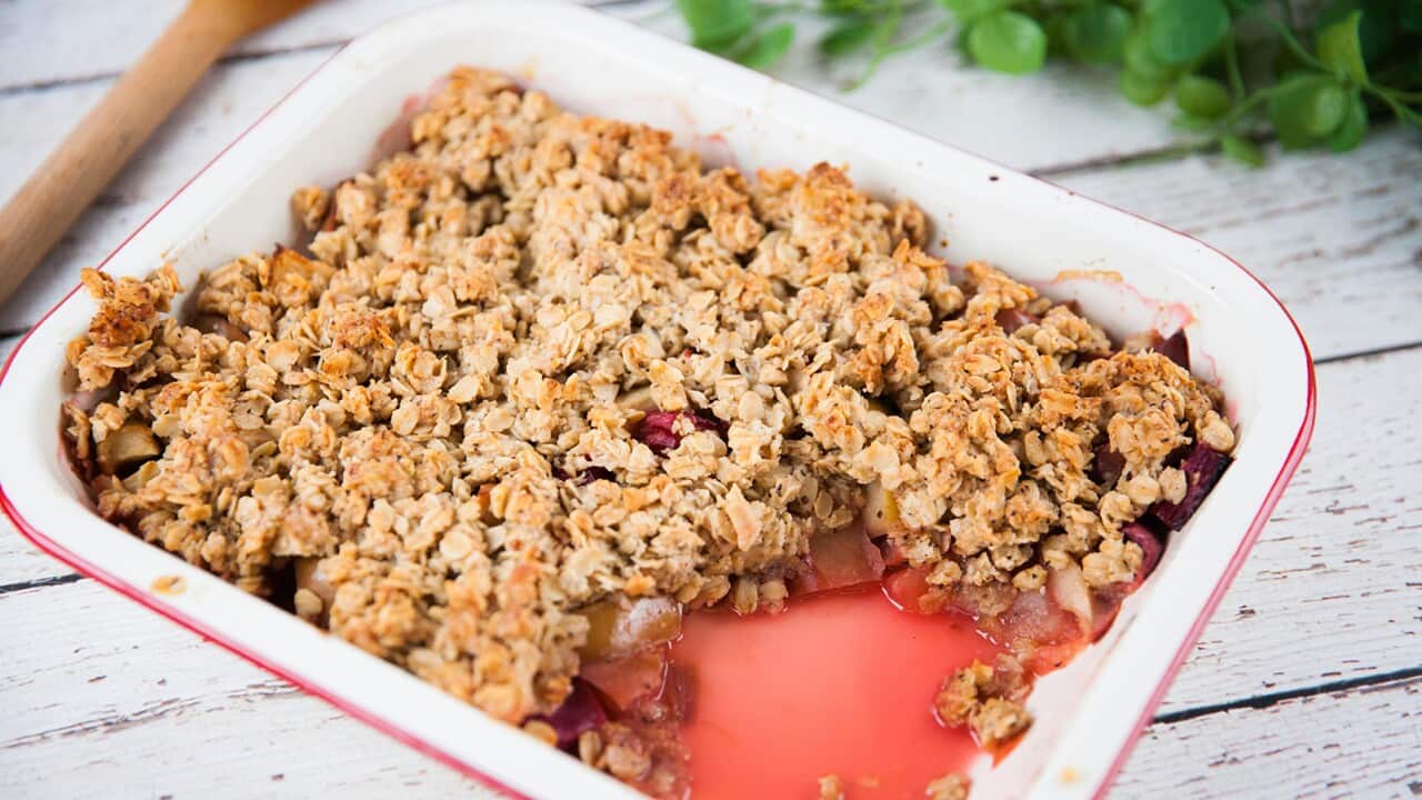 An old-style baking tray sits on a wooden surface. A portion of the crumble has been removed, showing the pink-tinged fruit under the chunky crumble topping.