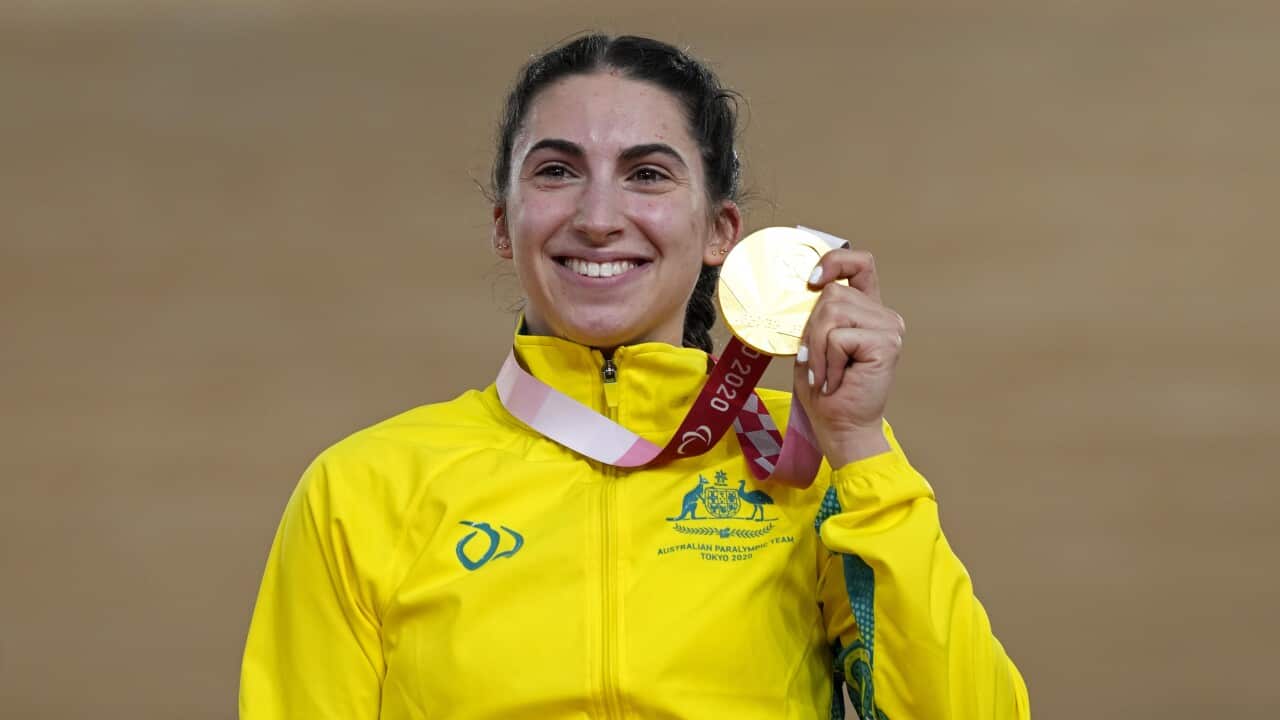 A young woman holding up a gold Paralympic medal. She is smiling and wearing a gold-coloured jacket with the Australian emblem on it.