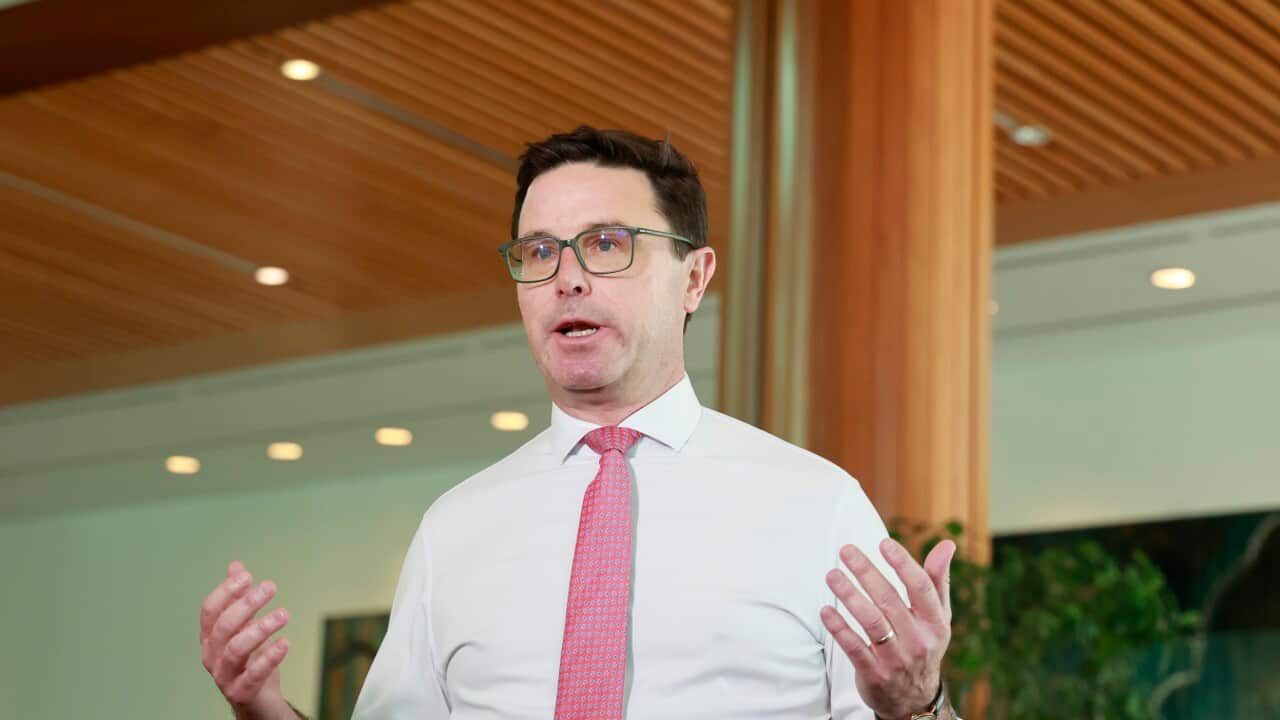 A middle-aged white man wearing a white shirt, red tie and glasses. His hands are slightly raised beside him as he speaks at a press conference in Parliament House in Canberra.