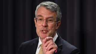 A man with grey hair, wearing glasses, a dark suit, which shirt, and yellow tie