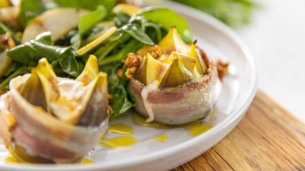 Two figs, cut open and wrapped in a band of pancetta, sit on a white plate, beside a pile of salad leaves.