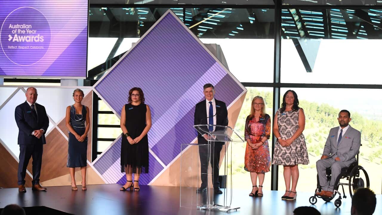 Seven individuals dressed in semi-formal attire are lined up on stage, with one man in a wheelchair.