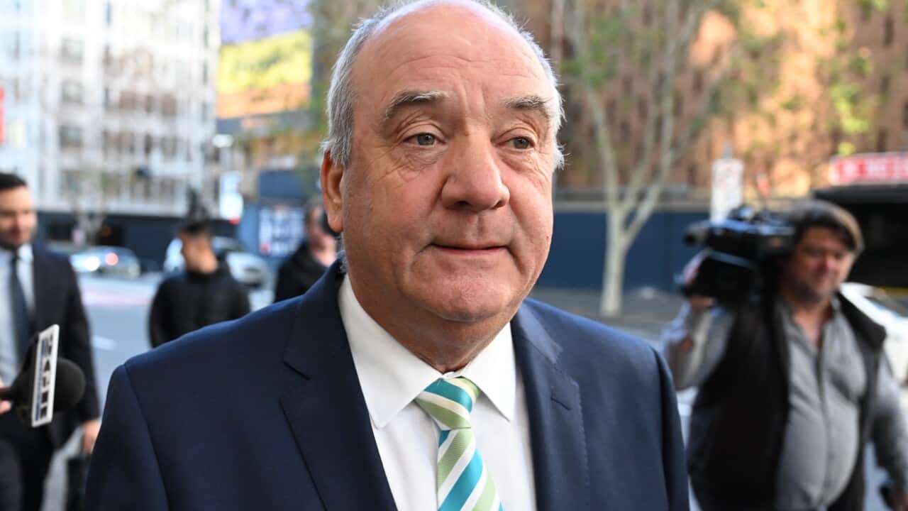 An elderly man in a blue suit and striped tie is walking down a street. There are camera operators and other people near him.