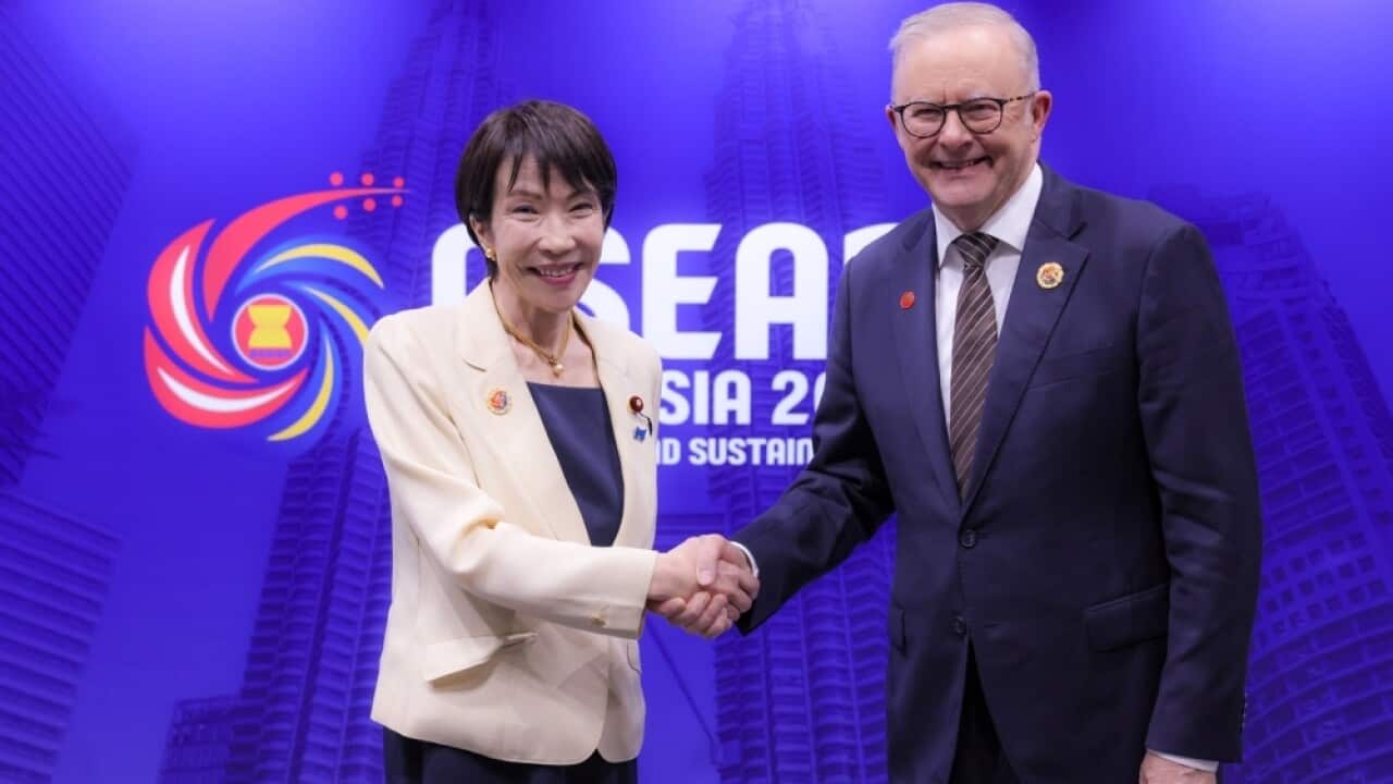 A man and a woman shaking hands, smiling at the camera in front of a blue wall