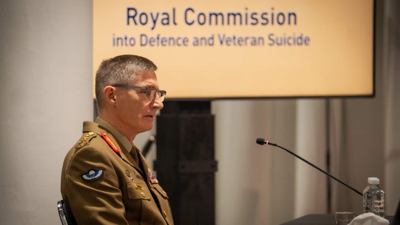 A man in a khaki military jacket speaks in front of a microphone