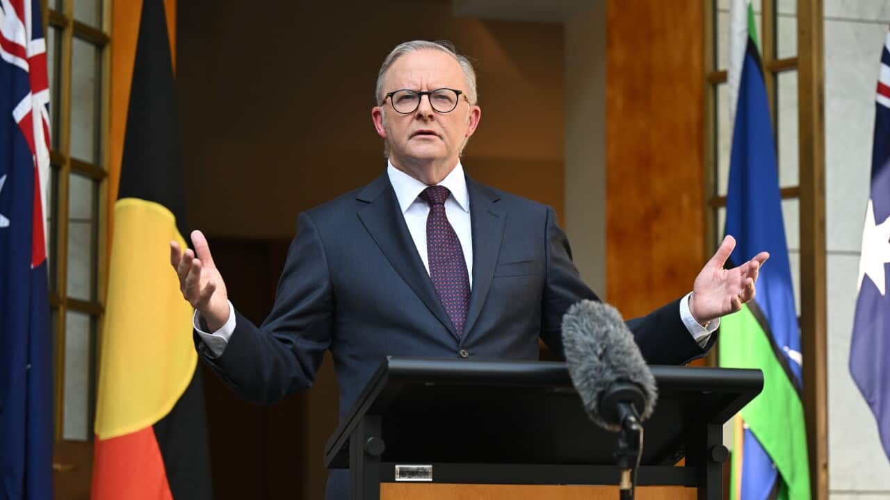 Anthony Albanese stands behind a lectern featuring the Commonwealth Coat of Arms, gesturing with open hands while flanked by the Australian, Aboriginal, and Torres Strait Islander flags.