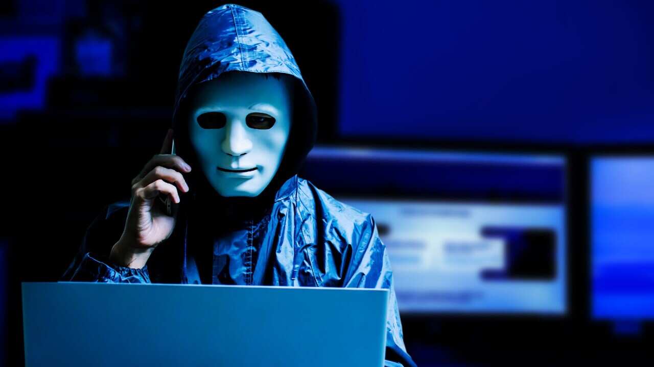 A Male Hacker Wearing Mask While Using Laptop At Table In Darkroom