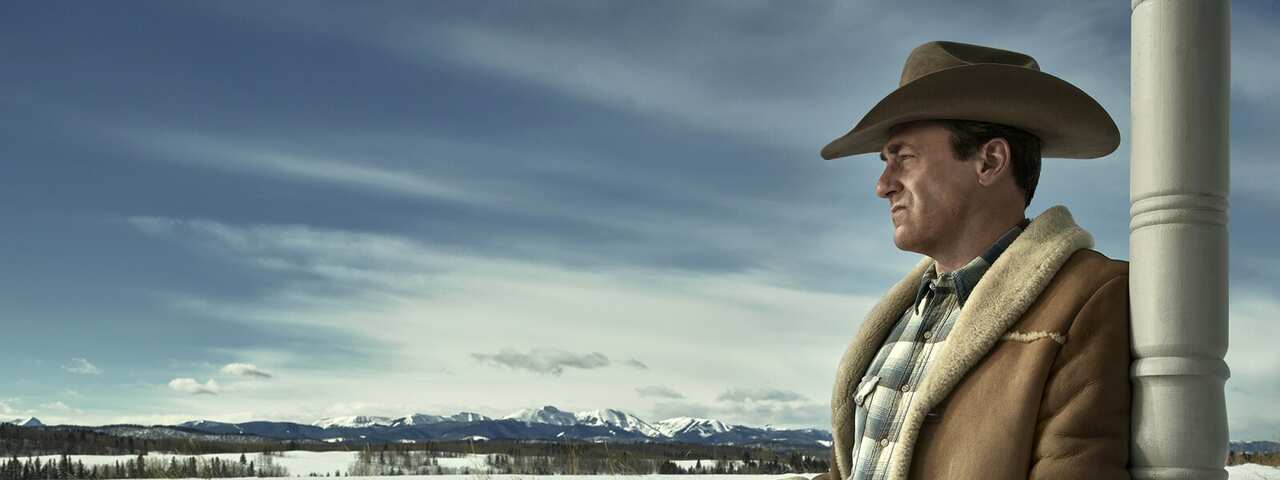 A man in a check shirt, thick jacket and western hat leans against a pole, side on, in front of a snowy landscape.