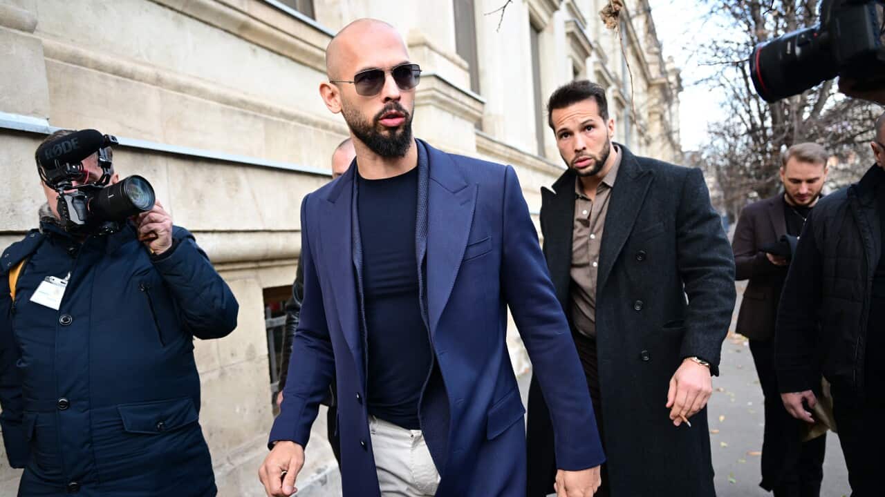 A man in a blue suit jacket walks next to a courthouse surrounded by reporters