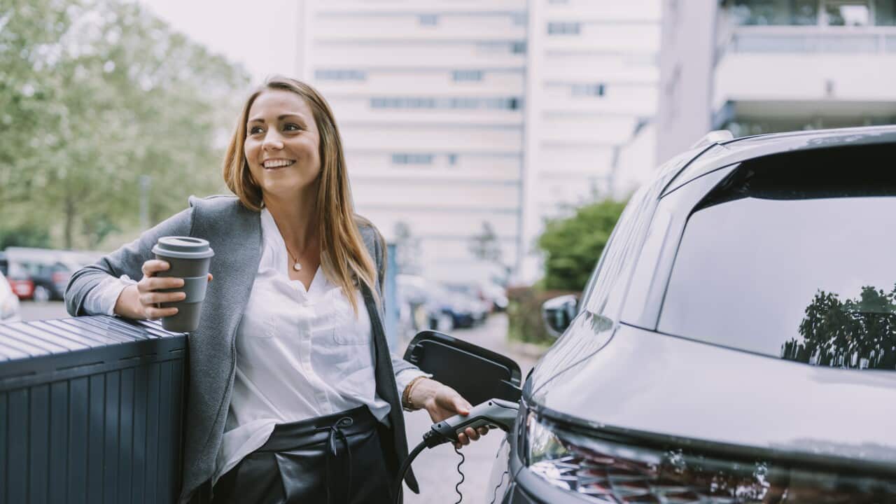 Happy woman with disposable coffee cup charging car at station