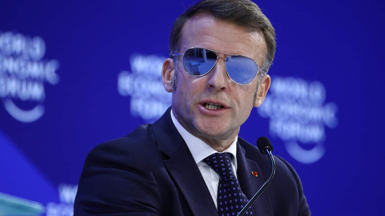 Emmanuel Macron wearing a dark suit and sunglasses and talking at a microphone