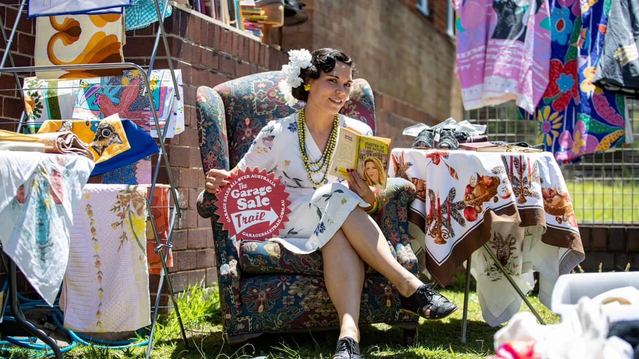 A woman in a vintage dress sits down on a sofa outdoors near vintage clothes diplayed on racks.