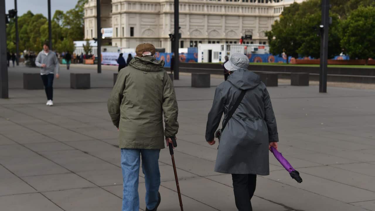 Two elderly people in coats walk away from the camera across a wide paved plaza, one using a cane and the other holding a purple umbrella, with a large ornate building in the background.