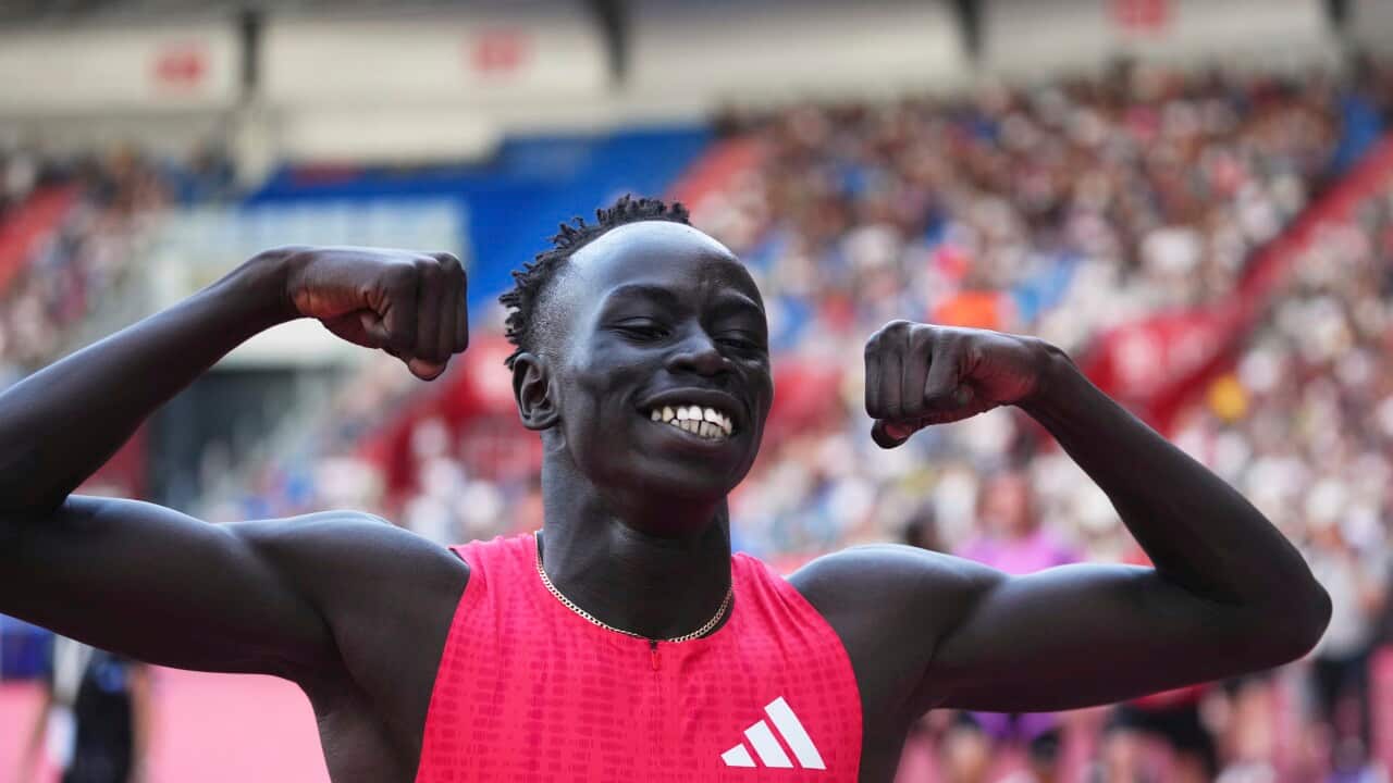 Gout Gout, in a red jersey with a white logo, smiles as he raises his arms in a sign of victory inside a stadium.