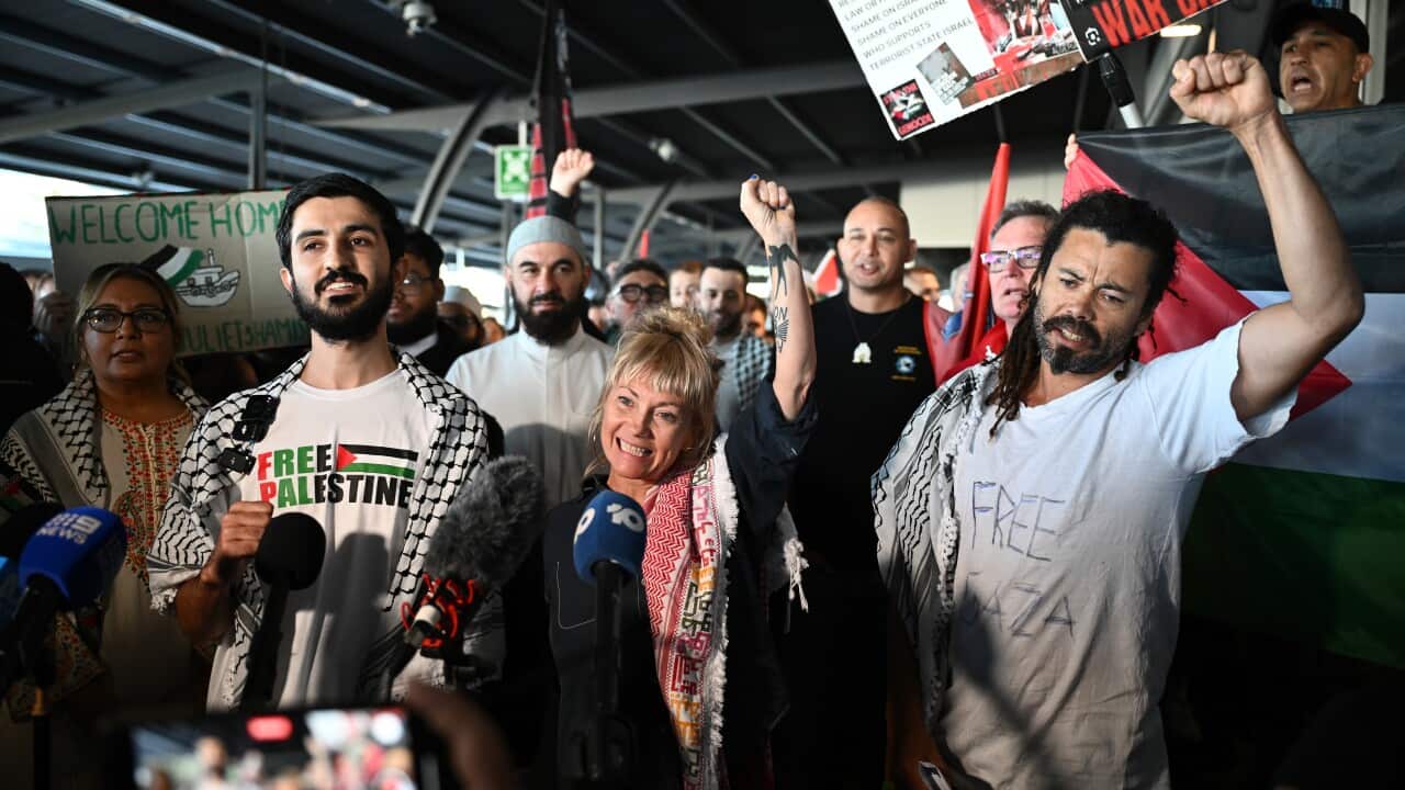 Three people smile and raise their fists as they stand in front of microphones. There's a crowd behind them, some holding posters.