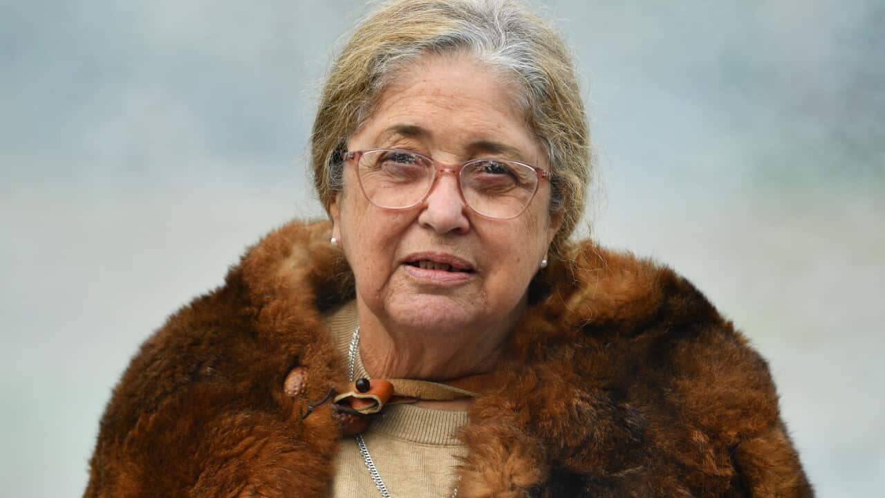 A woman in a fur coat addresses the media with a serious expression.