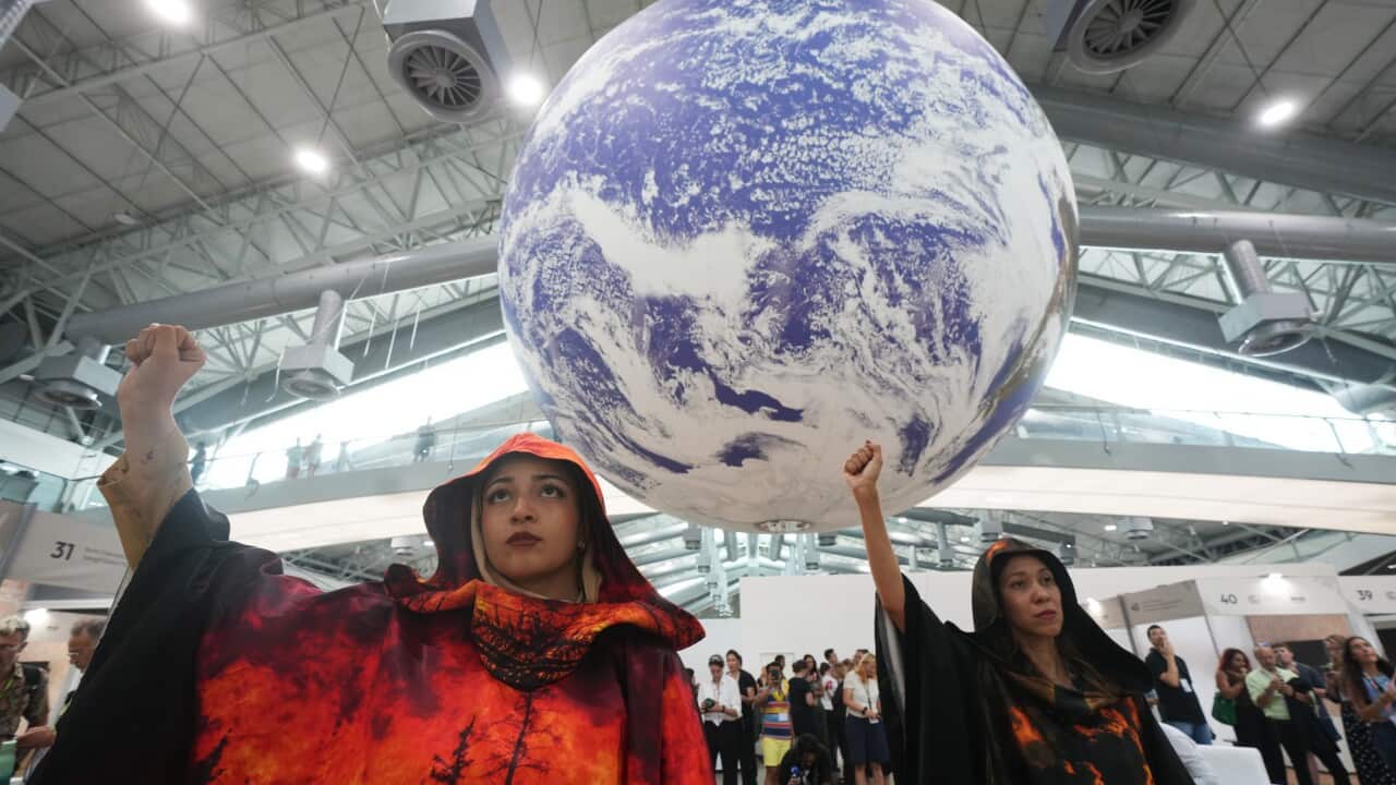 Two women in dark, hooded robes, one with a fiery red and black pattern and the other dark with an orange glow, stand indoors with their fists raised beneath a large, suspended inflatable globe.