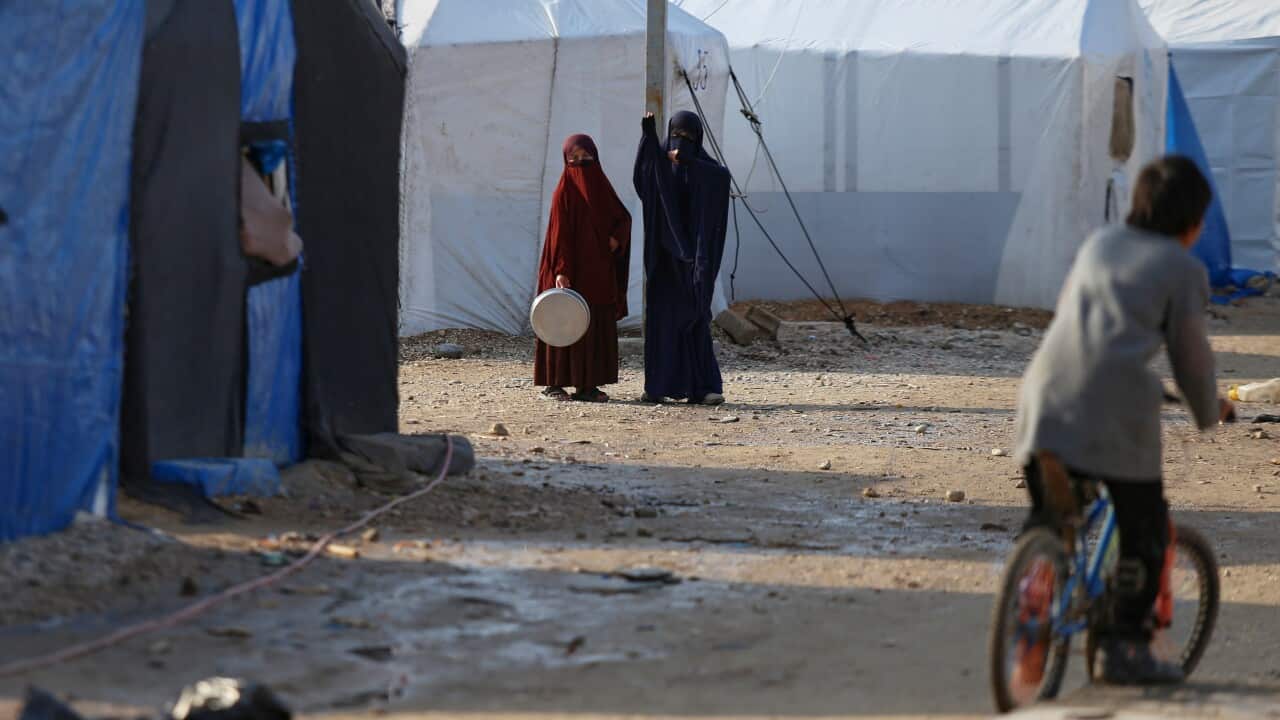 Two people wearing full-length niqabs, one in maroon and one in navy blue, stand between rows of makeshift tents in a camp. In the foreground, a child on a bicycle is blurred as they ride past.