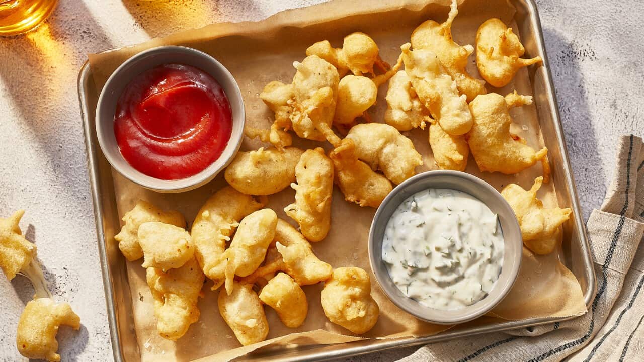 Little longish battered nuggets sit on baking paper on a metal tray. A bowl of sour cream and a bowl of red sauce also sit on the tray.