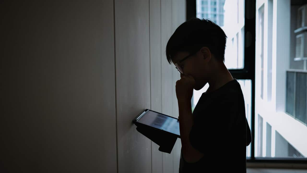 Silhouette photo of a young boy holding and looking at computer tablet against the wall inside an apartment alone.