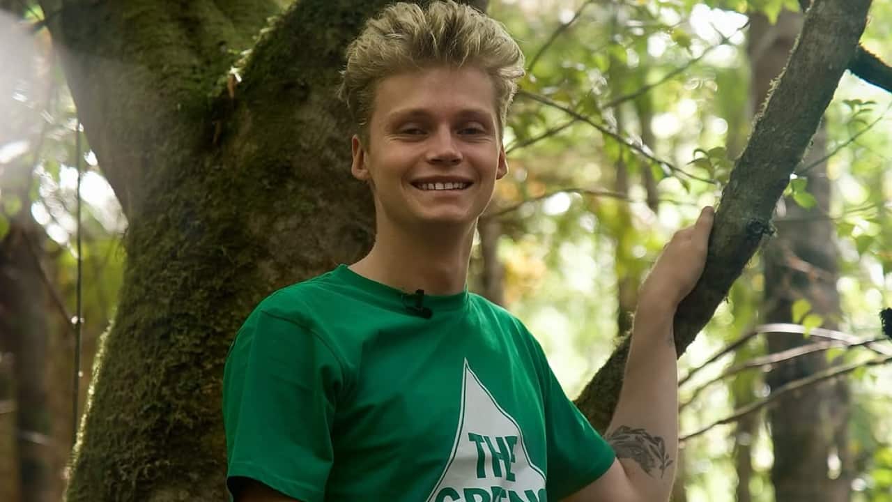 A young blonde male in a Greens party t-shirt leans against a tree.