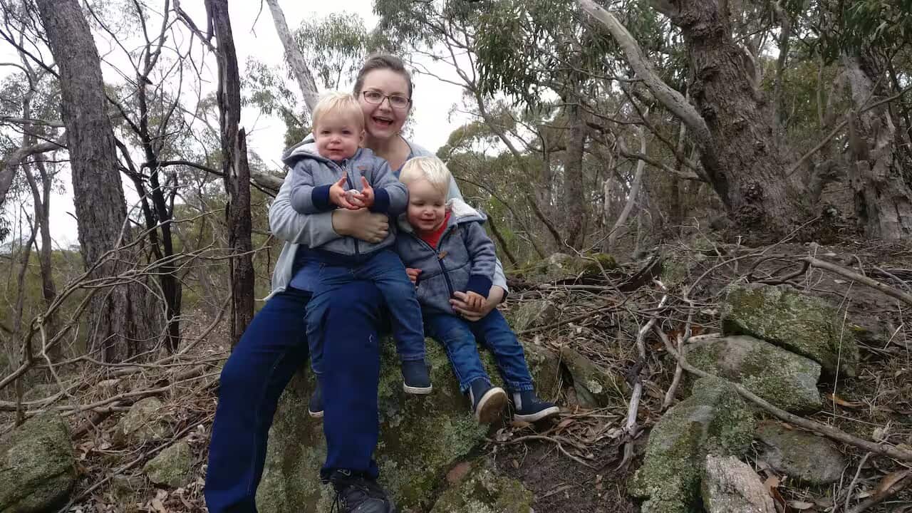 A middle-aged white woman holds her two young sons with blonde hair as they sit on a rock in the bush. 